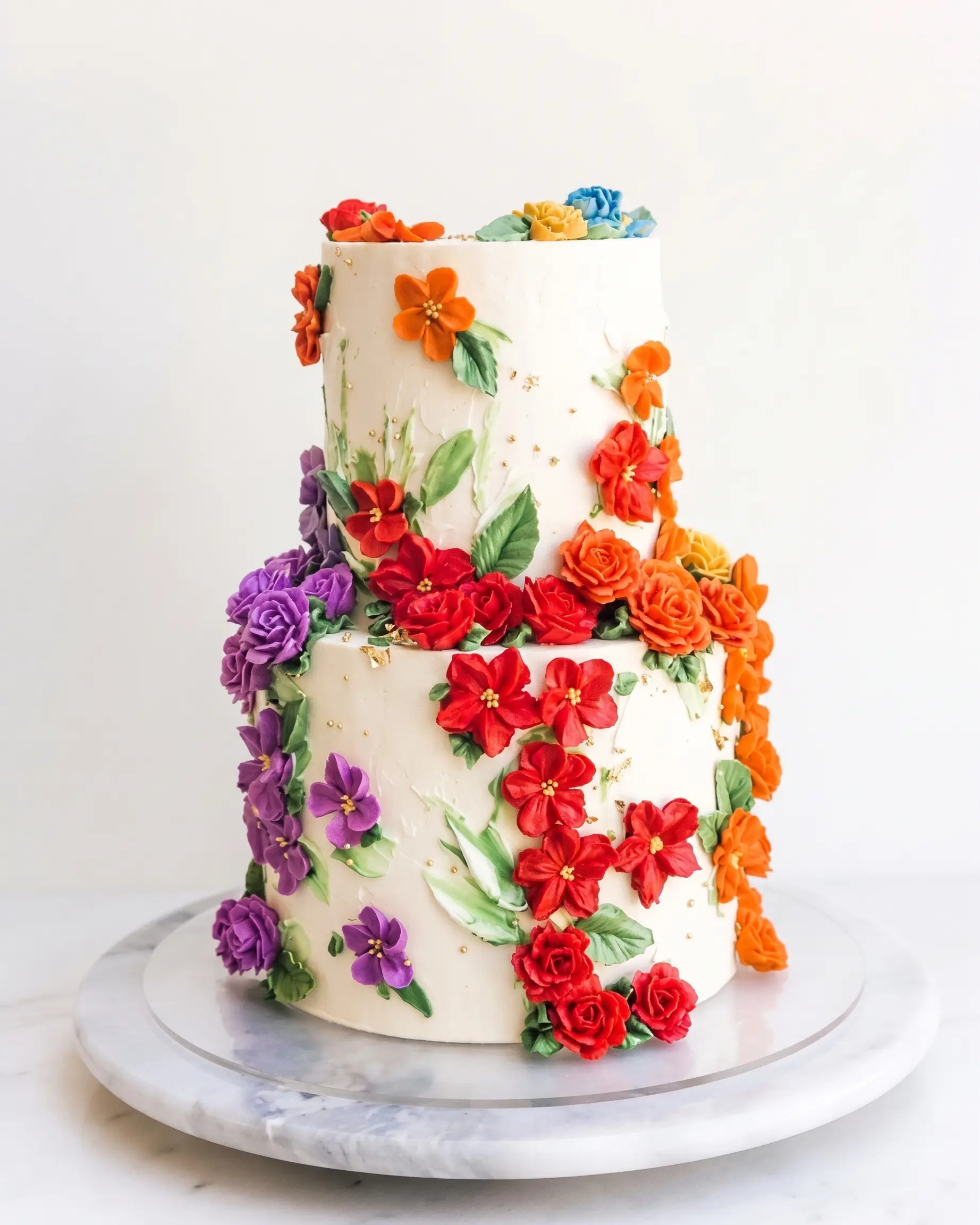 Two-tier white special event cake decorated with colourful sugar flowers, leaves and gold accents